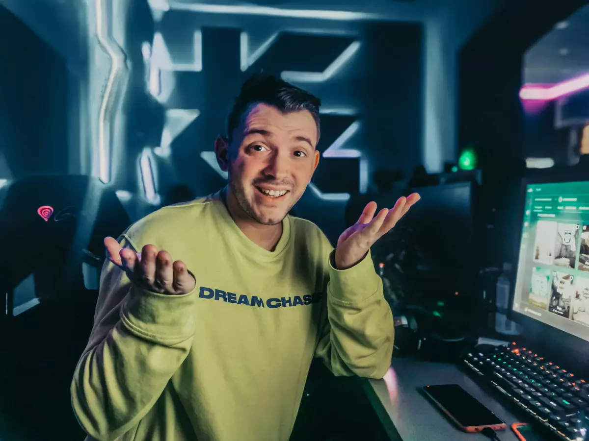 A young man in a yellow sweater sits at a desk in front of a computer, with a surprised expression on his face, looking at the camera. The background is a dark room with neon lights. 