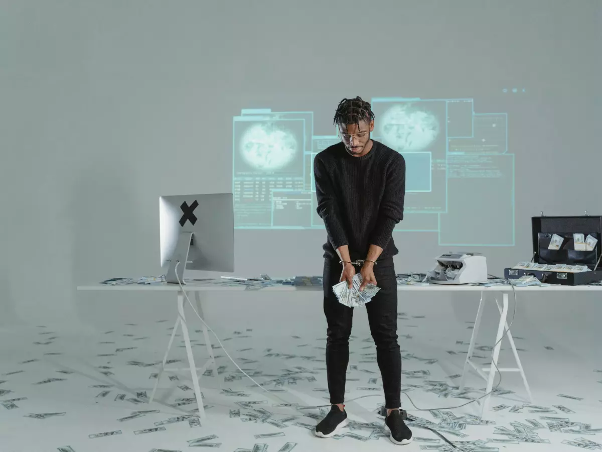 A man in a black shirt and pants is standing in a white room. In front of him is a table with some equipment, and he is looking towards the camera. In the background, a large screen is projecting images, likely of data.