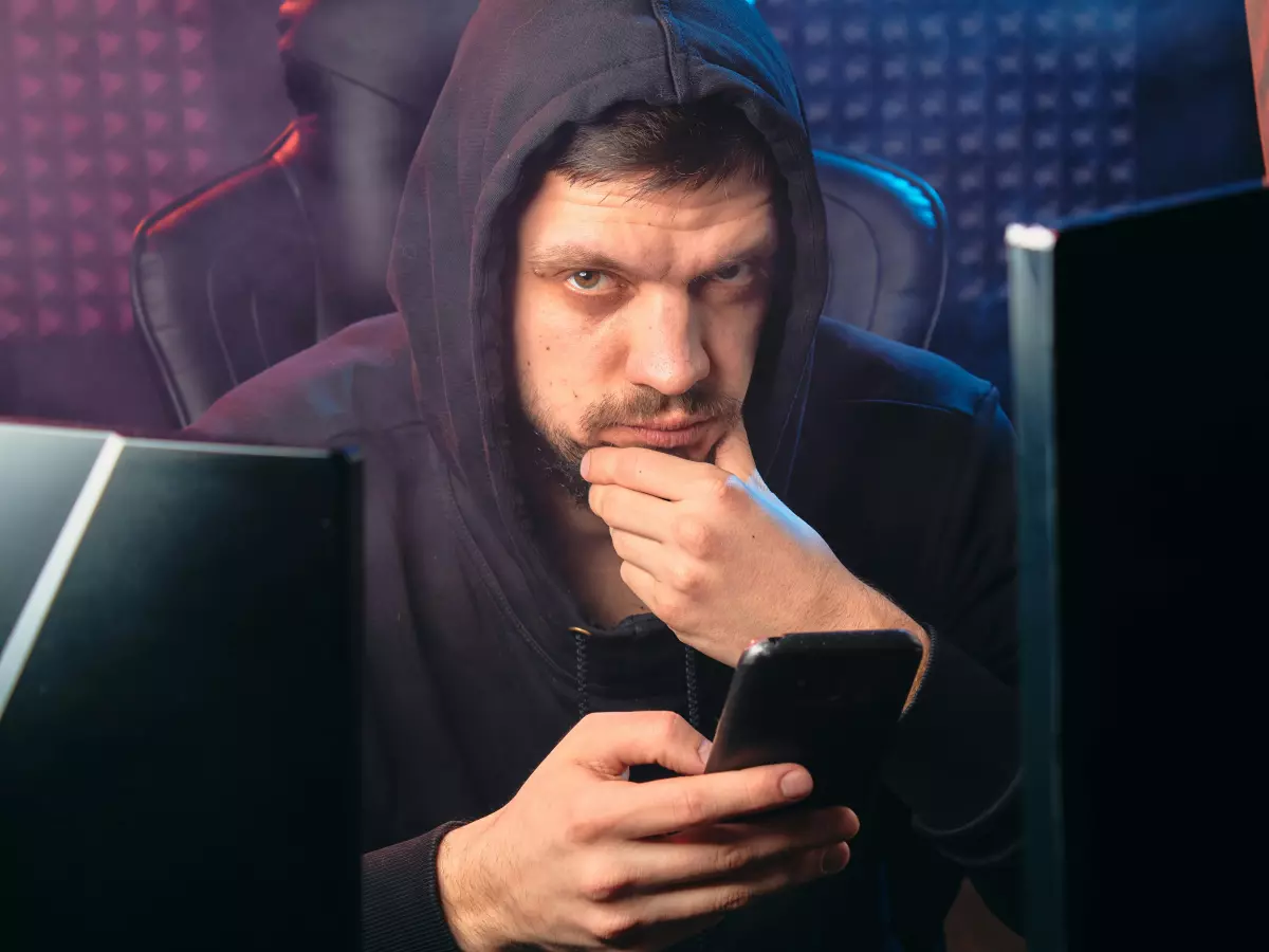 A hooded figure sits in a dimly lit room, staring intently at a smartphone screen. Two computer monitors are visible in the background, suggesting a hacking operation in progress.