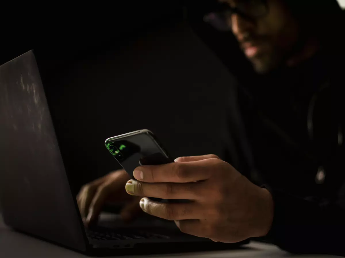 A person wearing a hoodie and glasses looking at a laptop screen. They are sitting at a desk and their face is obscured by the hood.