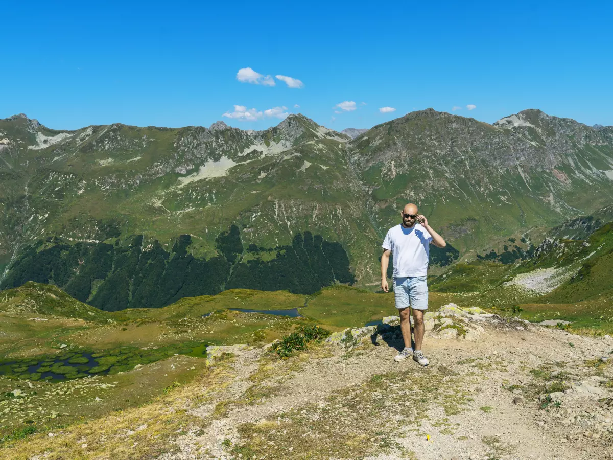 A man standing on a mountaintop, looking at his watch. The mountains and sky are in the background.