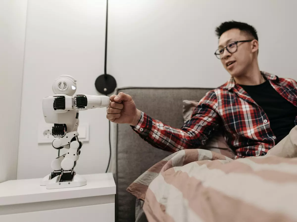 A man in a plaid shirt sits on a bed and reaches out his hand toward a robot that is standing on a nightstand. The robot is white and has a circular head with a camera on it.