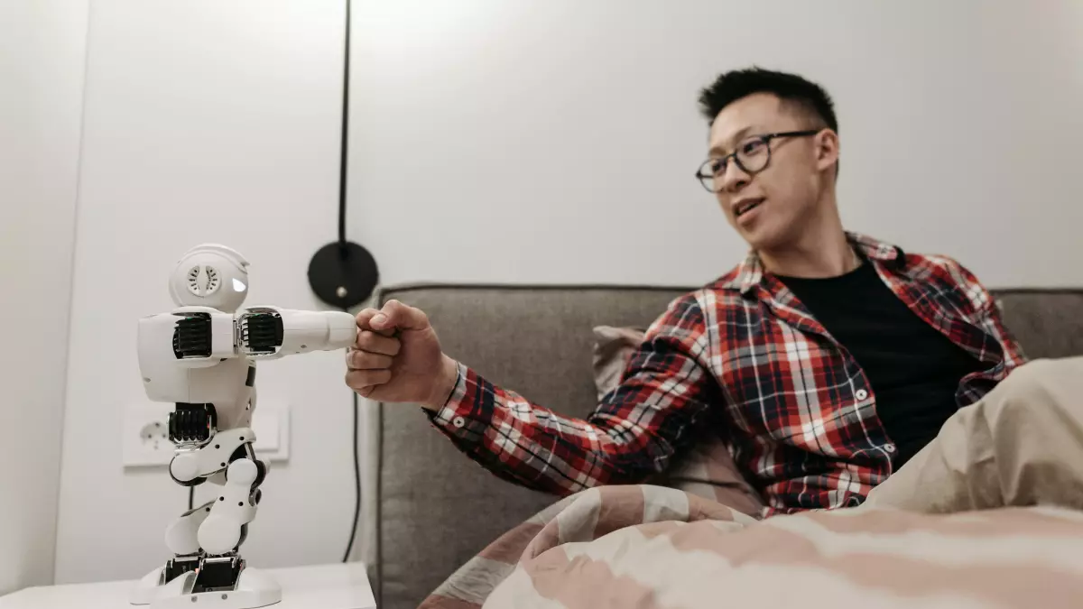 A man in a plaid shirt sits on a bed and reaches out his hand toward a robot that is standing on a nightstand. The robot is white and has a circular head with a camera on it.