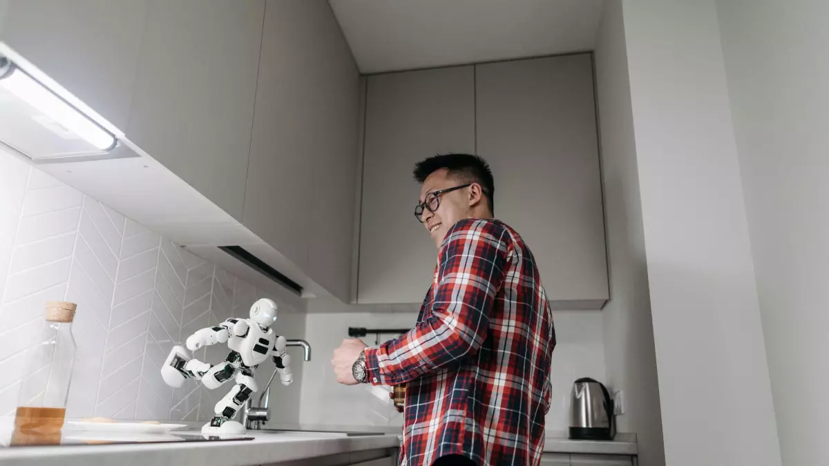 A man wearing a red and black checkered shirt and glasses stands in a kitchen, watching as a humanoid robot pours something into a cup.