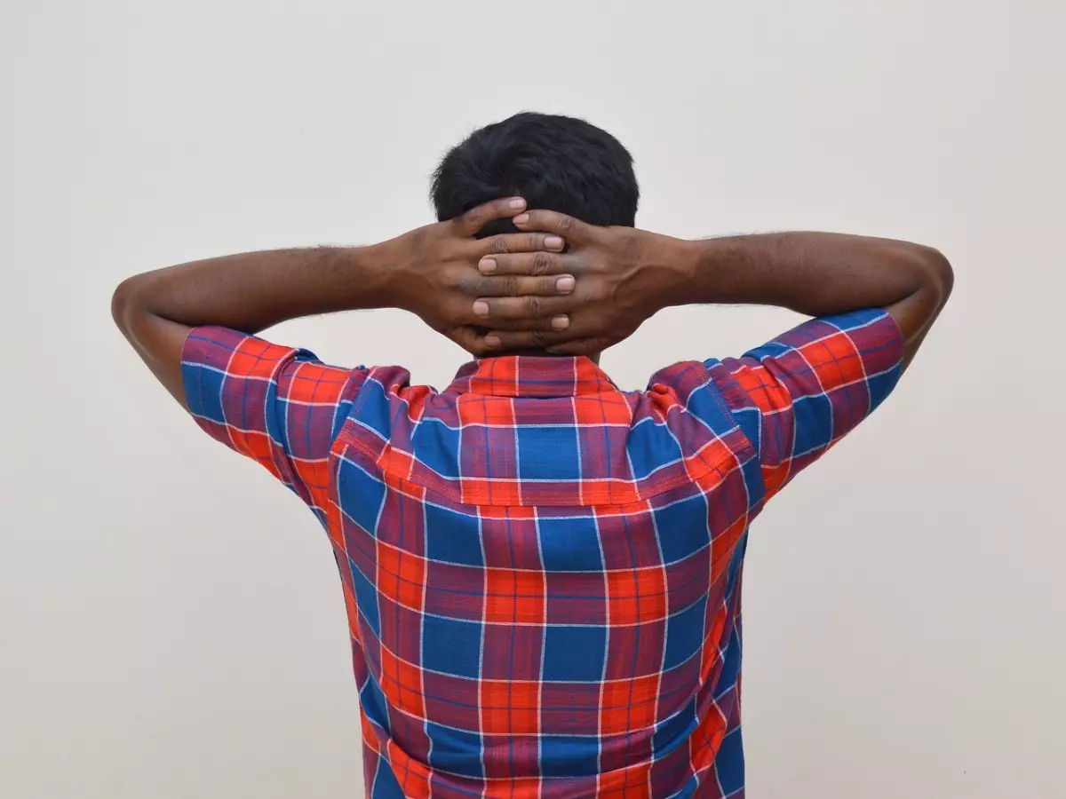 A man is standing with his arms behind his head and his head resting on his hands. He is wearing a plaid shirt and looks exhausted.