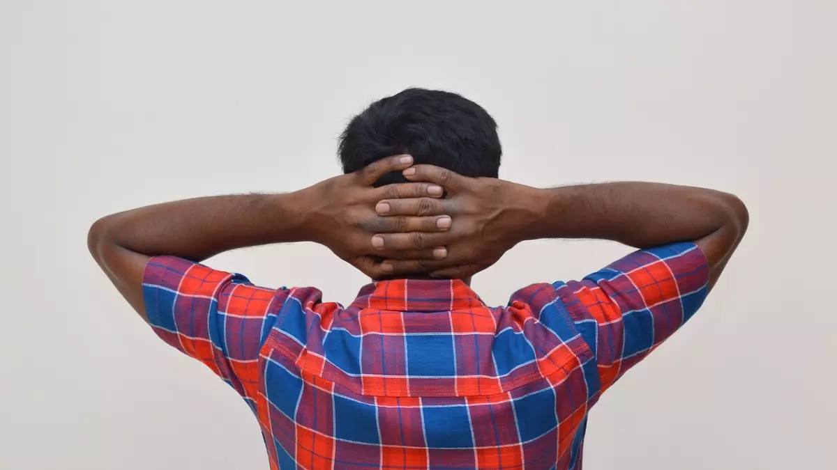 A man is standing with his arms behind his head and his head resting on his hands. He is wearing a plaid shirt and looks exhausted.