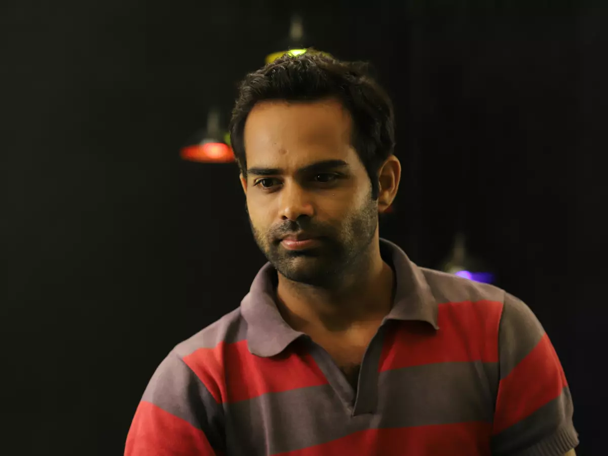 A man in a red and gray shirt, looking to the left, with a dark background and some blurred colored lights