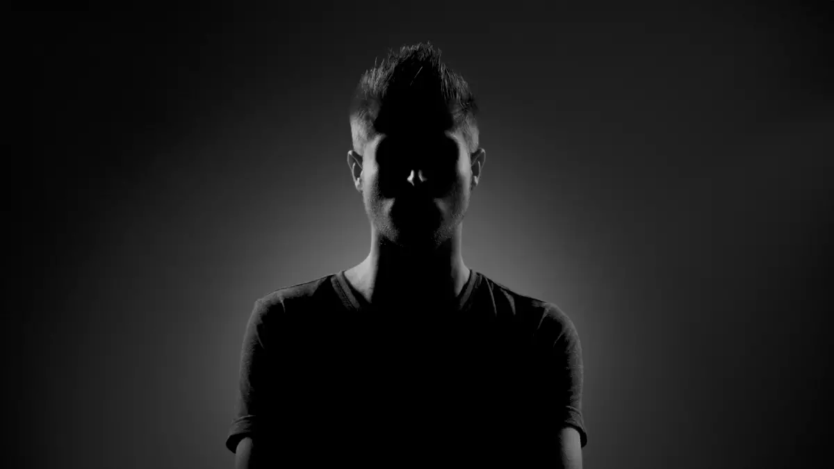 A man standing in front of a dark background, his face partially obscured by shadows.