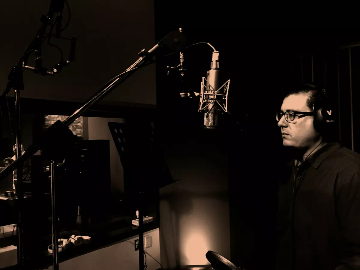 A man in a recording studio, singing into a microphone with headphones on. The image is in sepia tone.