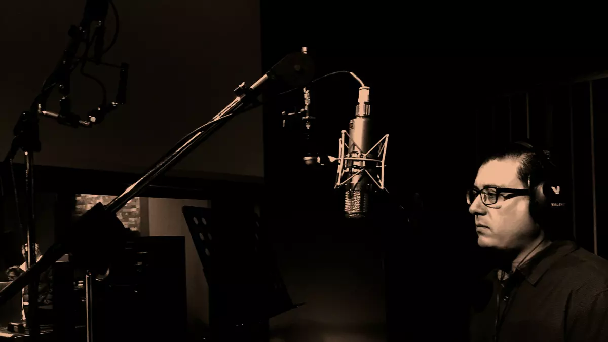 A man in a recording studio, singing into a microphone with headphones on. The image is in sepia tone.