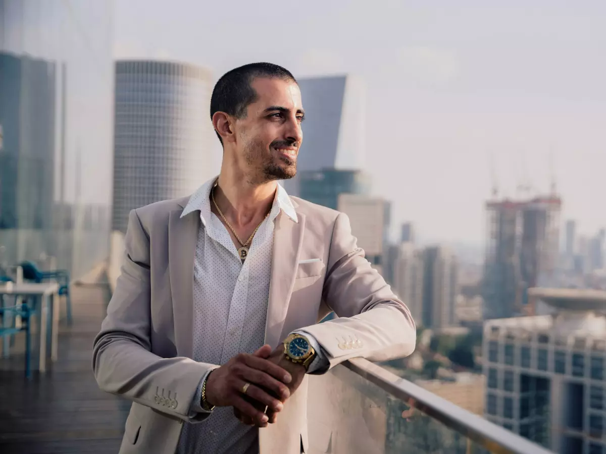 A man in a suit stands on a balcony overlooking a city skyline. He is looking to the right with a smile on his face.