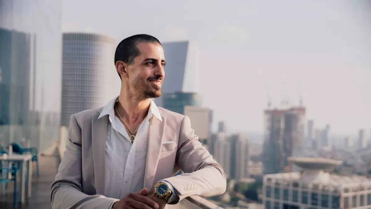 A man in a suit stands on a balcony overlooking a city skyline. He is looking to the right with a smile on his face.