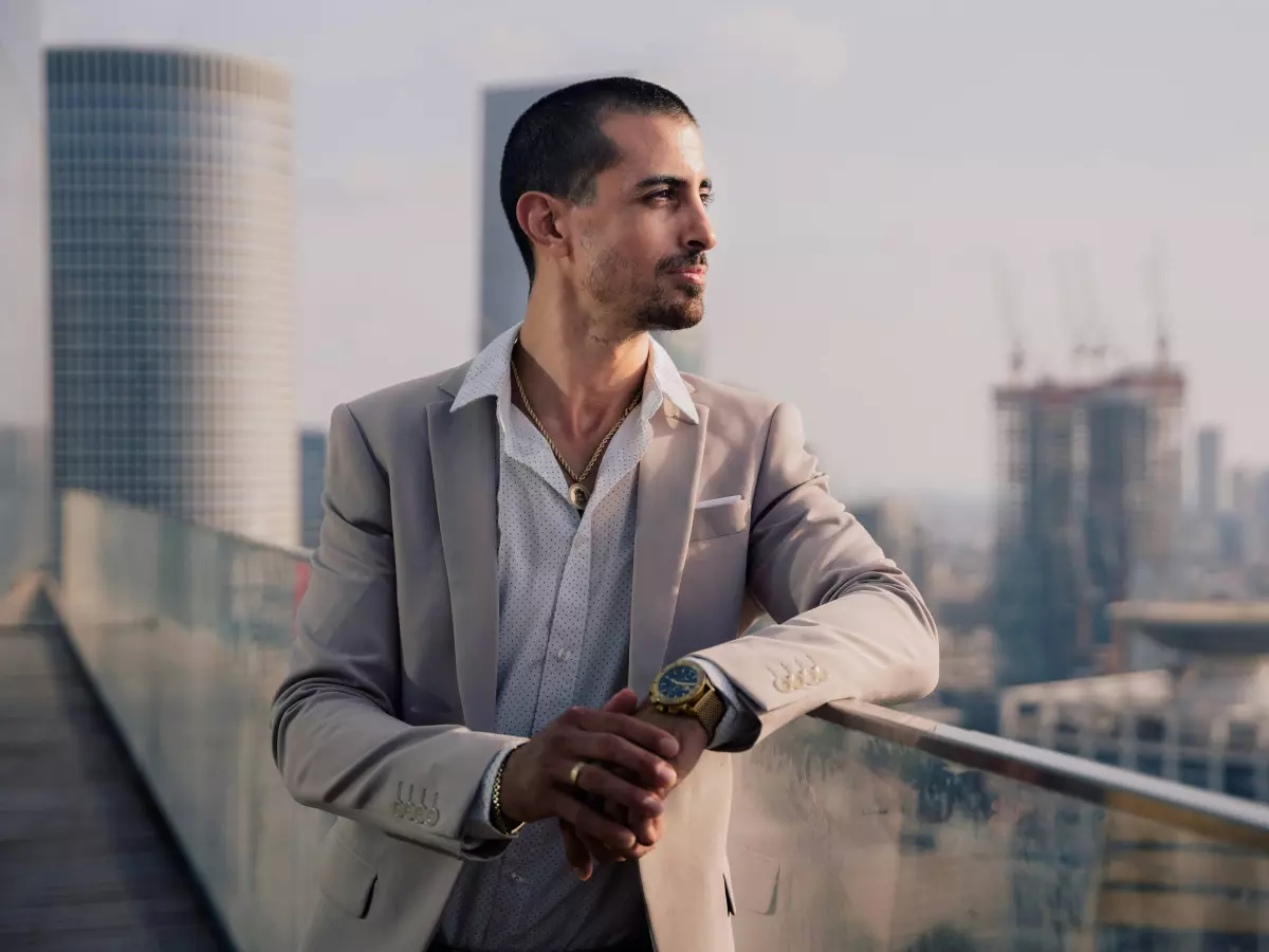 A young man in a suit is standing on a rooftop, looking out over a cityscape. He is wearing a gold watch and has a thoughtful expression on his face.