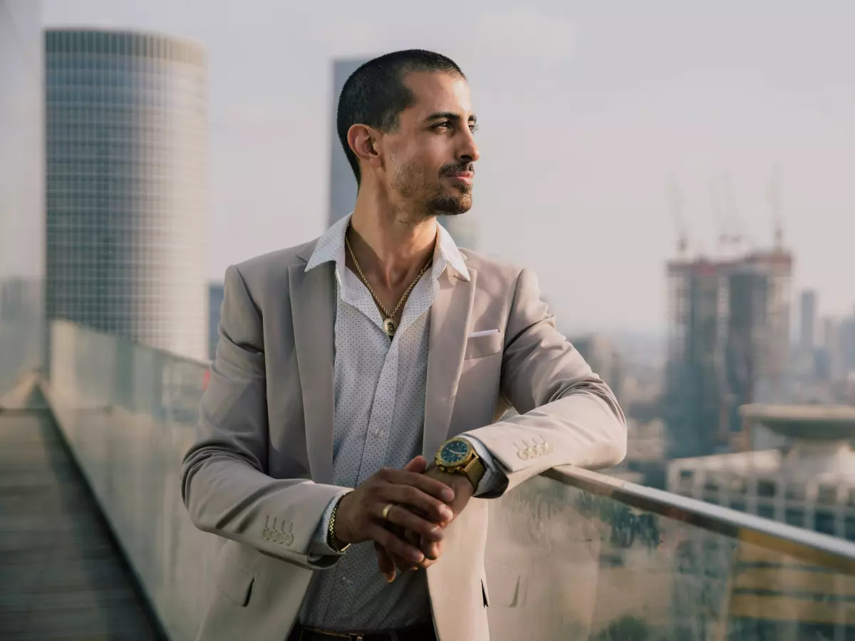 A man in a suit is standing on a balcony overlooking a city skyline. He is looking off to the side and appears to be deep in thought.