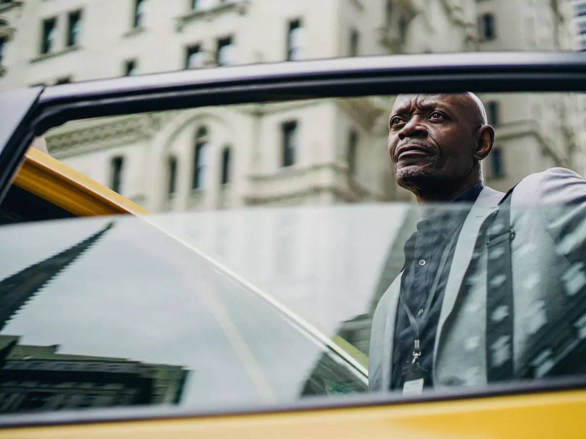 A man in a suit is sitting in the back of a yellow taxi, looking out the window at the city.