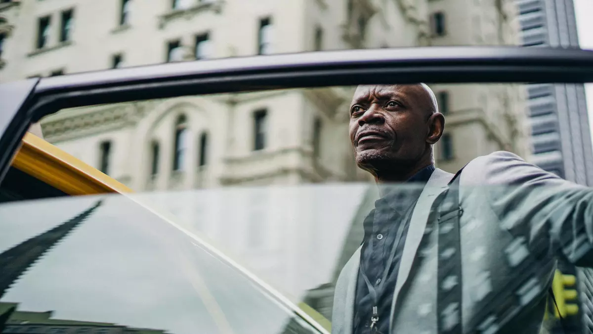 A man in a suit is sitting in the back of a yellow taxi, looking out the window at the city.