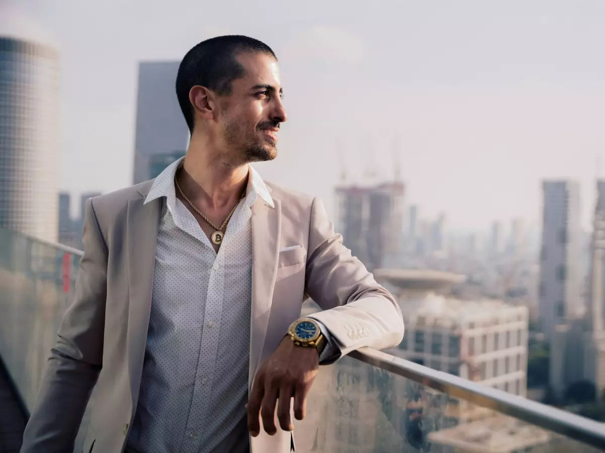 A man in a suit stands on a rooftop overlooking a city skyline. He is wearing a watch and a gold chain necklace.