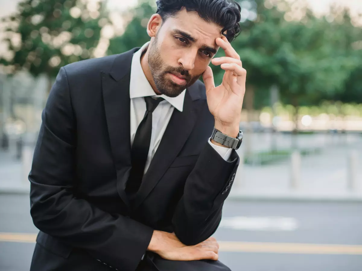 A man in a suit sits outside, holding his head in his hand, looking troubled.