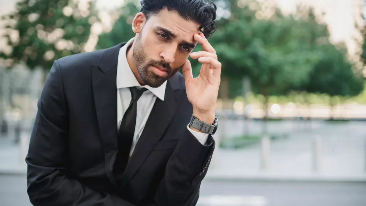 A man in a suit sits outside, holding his head in his hand, looking troubled.