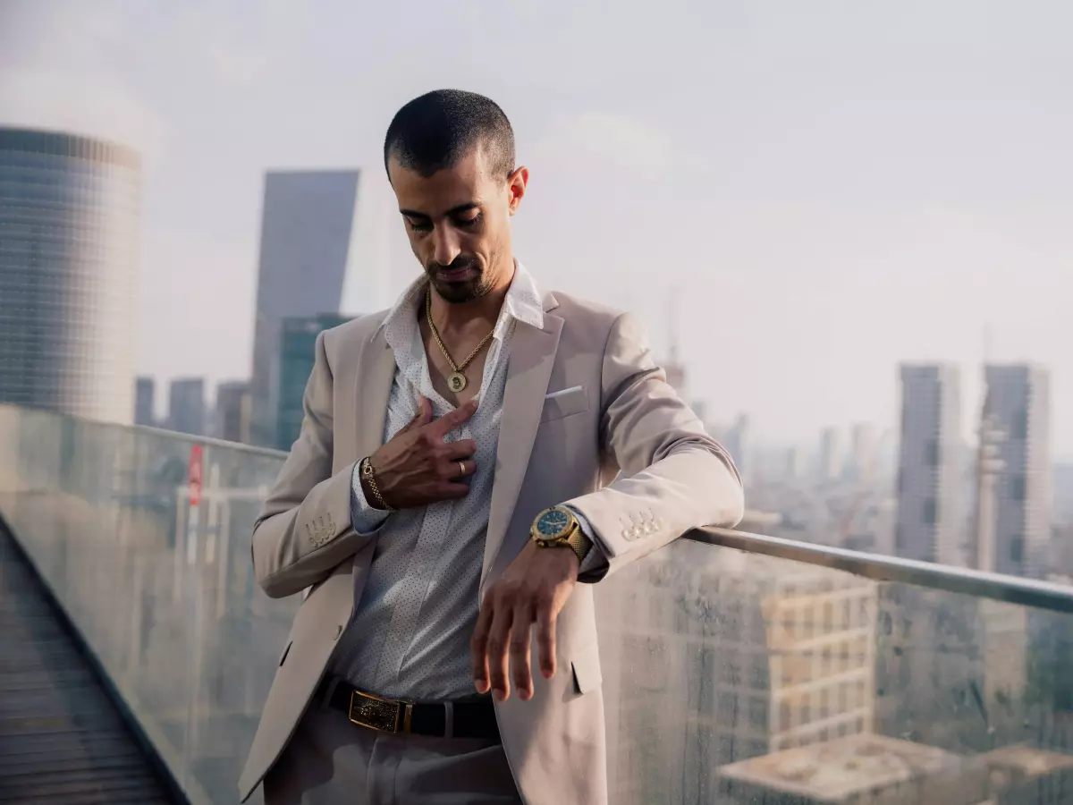 A man in a suit and a gold watch looks at his watch on a rooftop with city buildings in the background. 