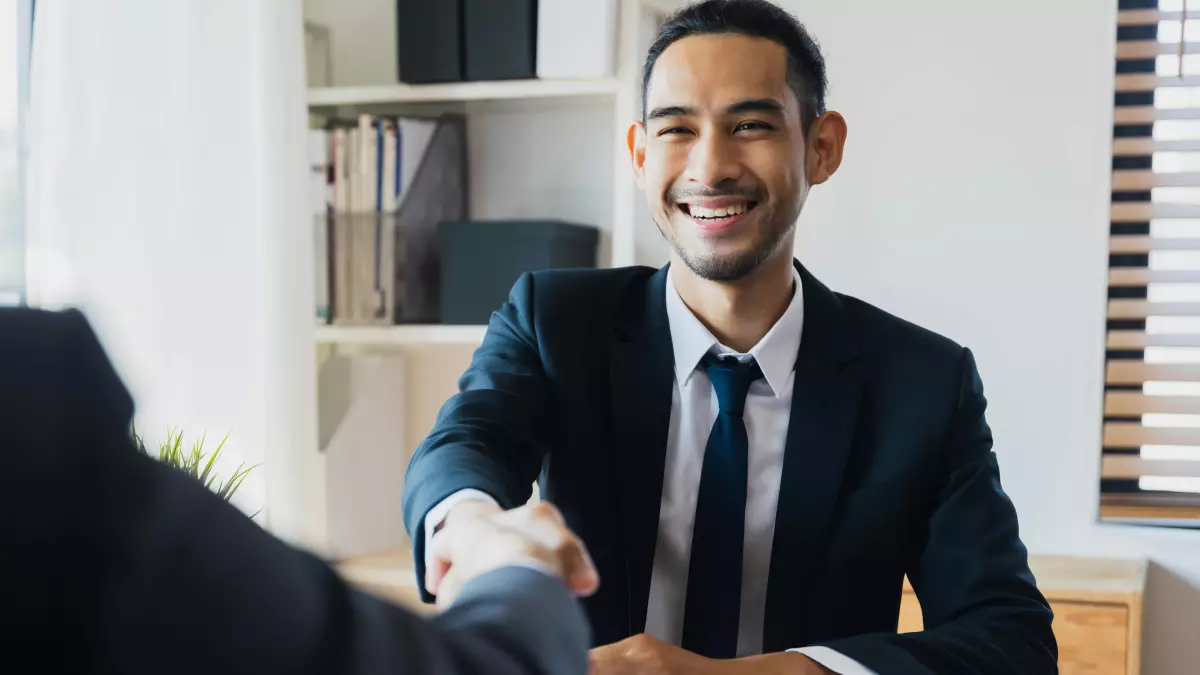 A man in a suit shakes hands with another man in a suit. They are both smiling.