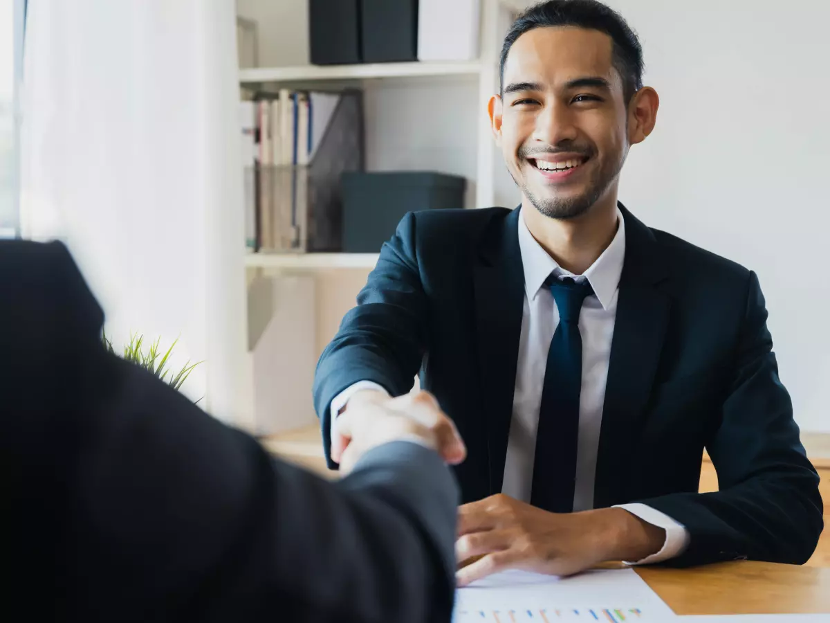 A man in a suit shakes hands with another man in a suit. They are both smiling.