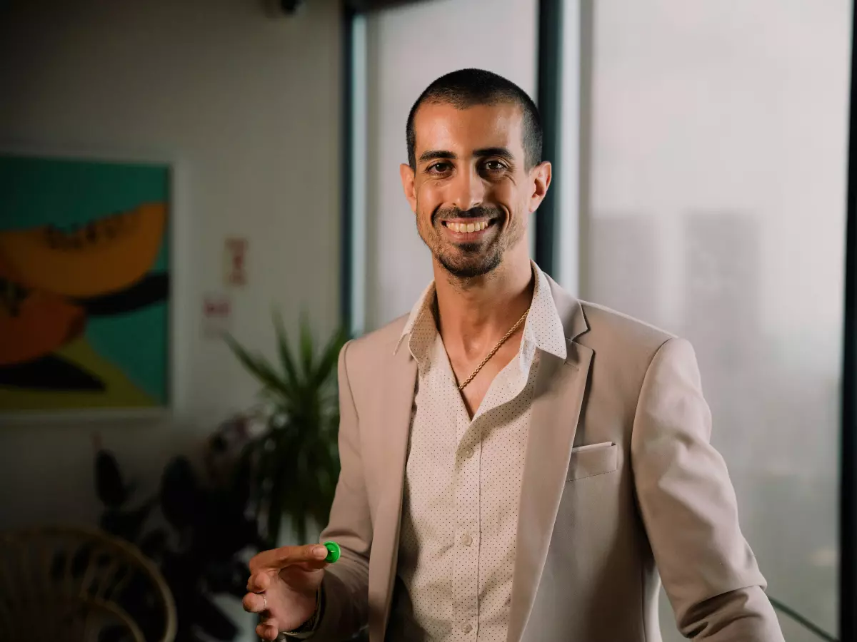 A man in a light brown blazer is smiling at the camera, standing in a well-lit office space.