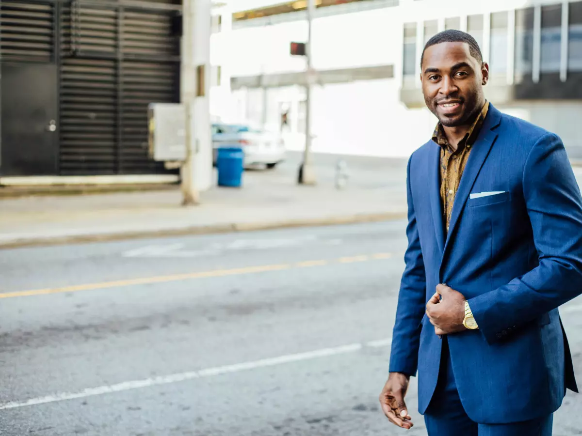 A man in a suit is standing in front of a city background. He is looking at the camera. 