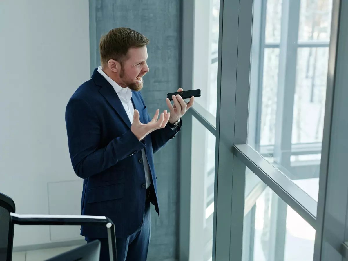 A man in a suit stands near a window, holding his phone and gesturing with frustration.