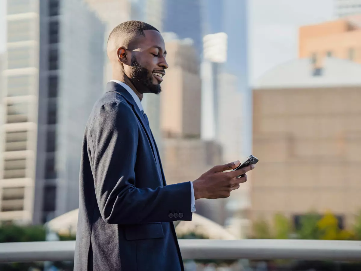 A man in a suit stands on a rooftop and is using his smartphone. The city skyline is visible behind him.