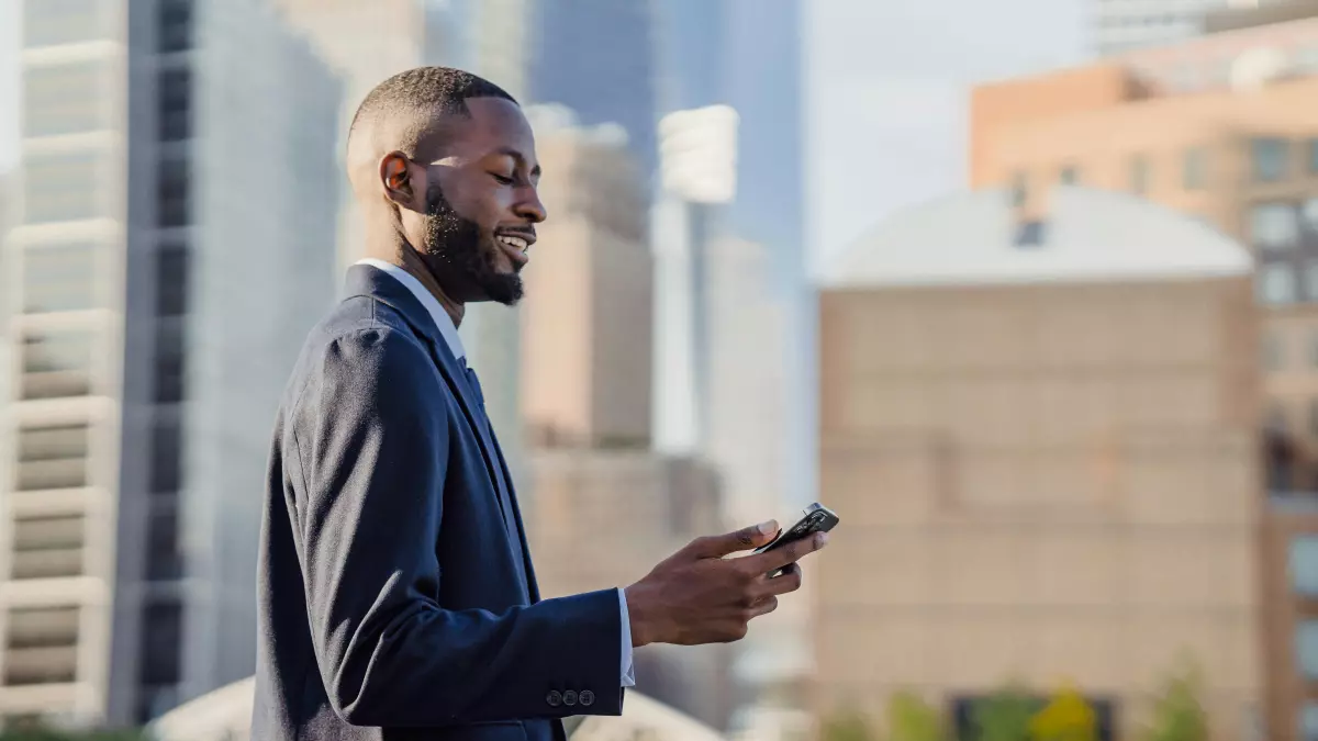 A man in a suit stands on a rooftop and is using his smartphone. The city skyline is visible behind him.