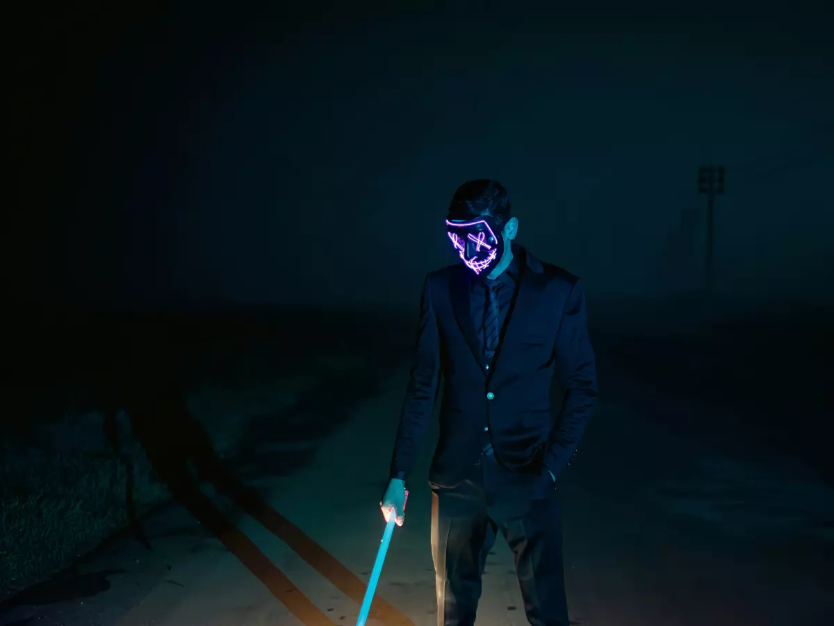 A man wearing a mask standing alone in a foggy, dark setting. He is wearing a suit and holding a glowing stick. 