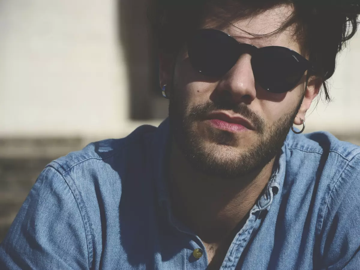 A man wearing sunglasses and a denim shirt is sitting on a staircase, looking down.