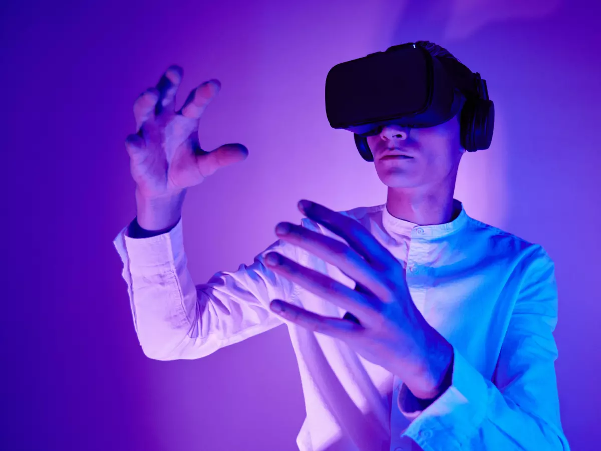 A man is wearing a virtual reality headset and is interacting with the virtual world with his hands. The room is dimly lit with purple light.