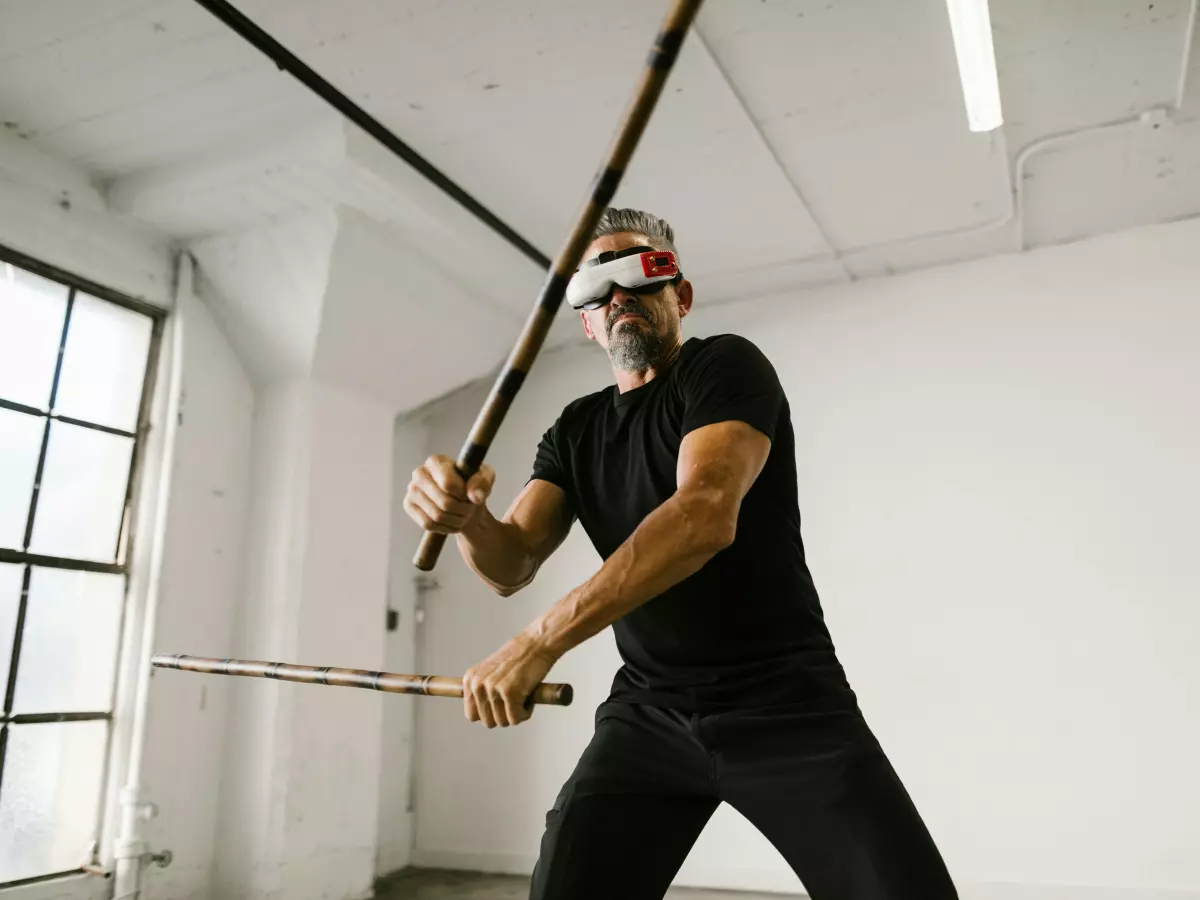 A man wearing a VR headset is holding two sticks, one in each hand, and is moving his arms around. He is wearing a black tank top and black pants. The background is a white wall with some pipes running along the top. 