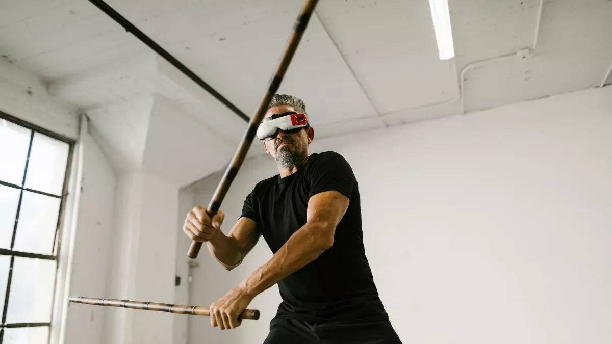 A man wearing a VR headset is holding two sticks, one in each hand, and is moving his arms around. He is wearing a black tank top and black pants. The background is a white wall with some pipes running along the top. 