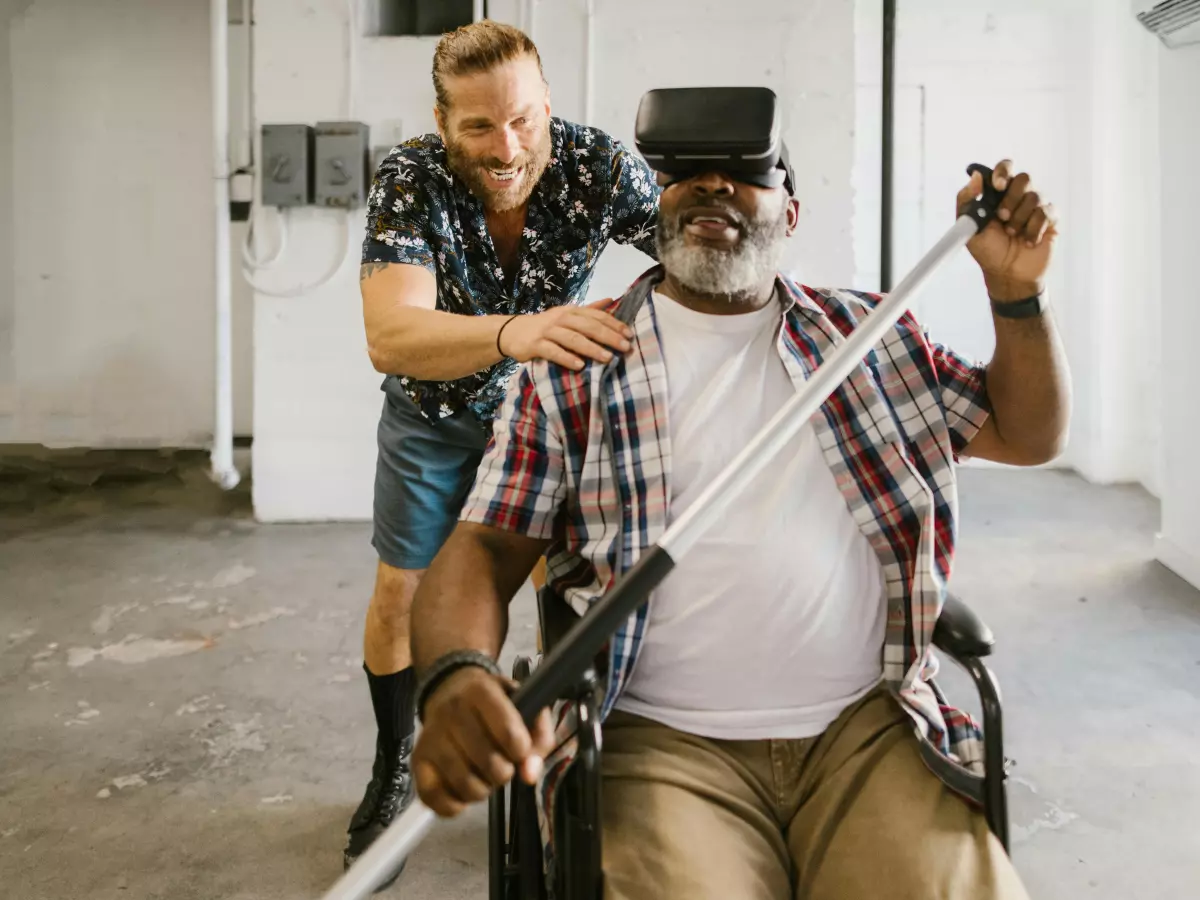 A man in a wheelchair is wearing a VR headset and being assisted by another man.