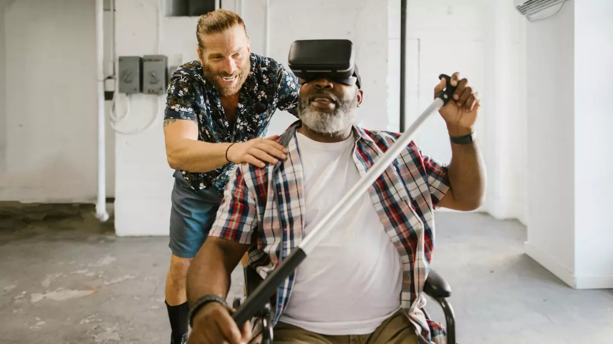 A man in a wheelchair is wearing a VR headset and being assisted by another man.