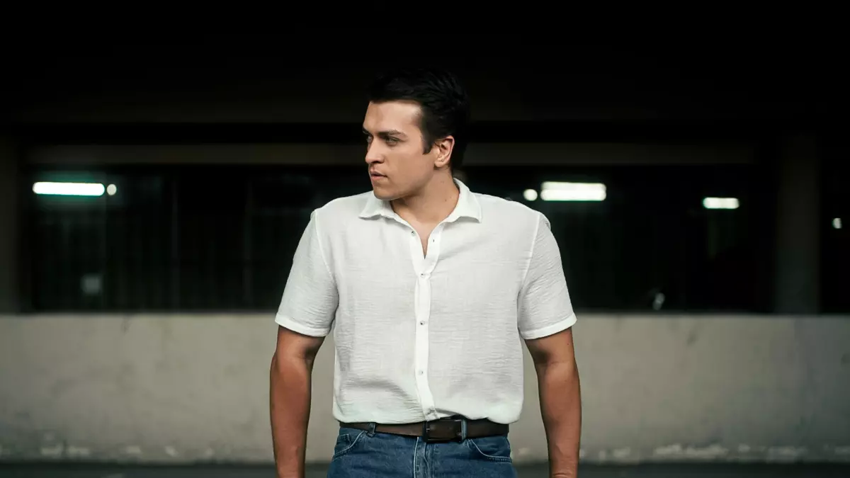 A man wearing a white shirt and blue jeans stands in a dimly lit environment, his gaze fixed on something beyond the frame. 