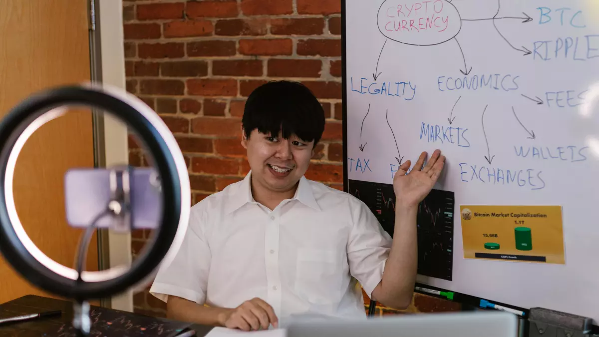 A man in a white shirt is sitting at a desk in front of a whiteboard with diagrams on it. He is pointing at the whiteboard with his right hand and smiling. There is a ring light and a microphone in front of him.