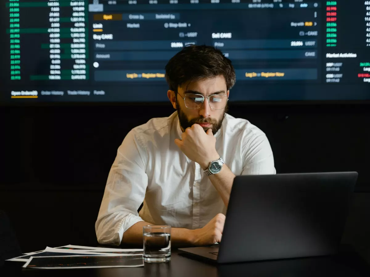 A young man in a white shirt and glasses sits at a desk with a laptop and a glass of water. Behind him, a large screen displays a stock market graph.