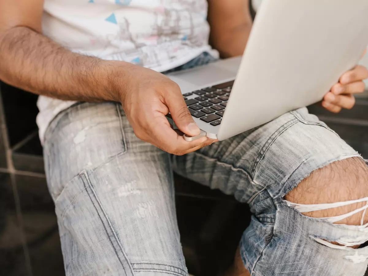 A close-up of a person's hands typing on a silver MacBook laptop. The person is wearing jeans and a white t-shirt. The background is blurred.