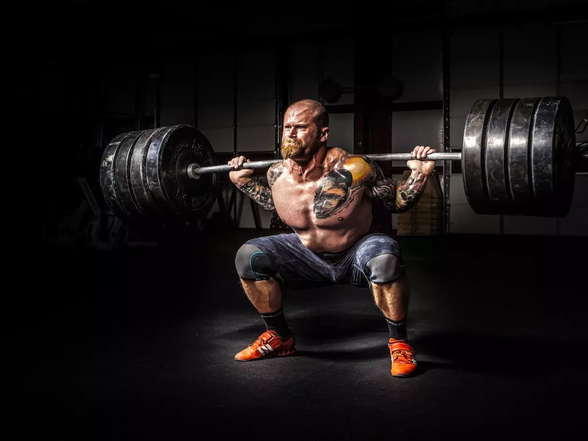 A man is lifting a heavy weight in a gym. He is muscular and has a beard. The image is dramatic and conveys strength and power.