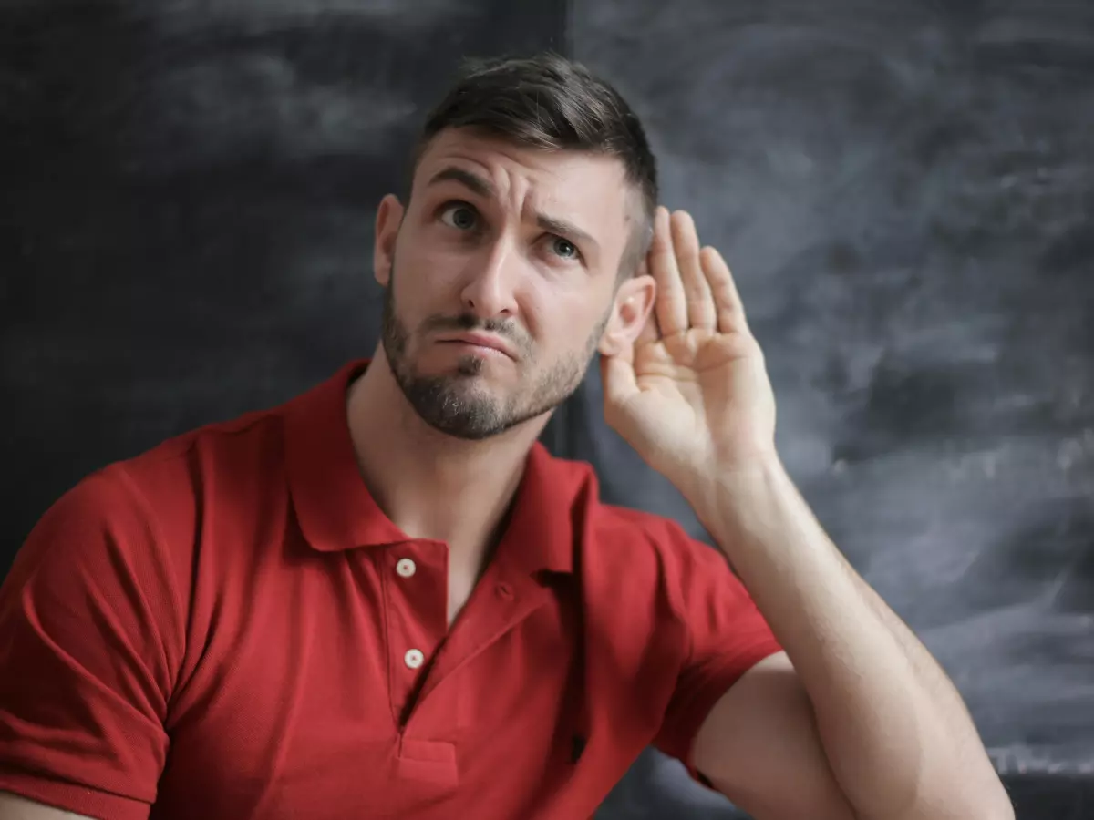 A man with short brown hair is wearing a red polo shirt and is listening intently. He is holding his hand to his ear, seemingly curious to hear what is being said.