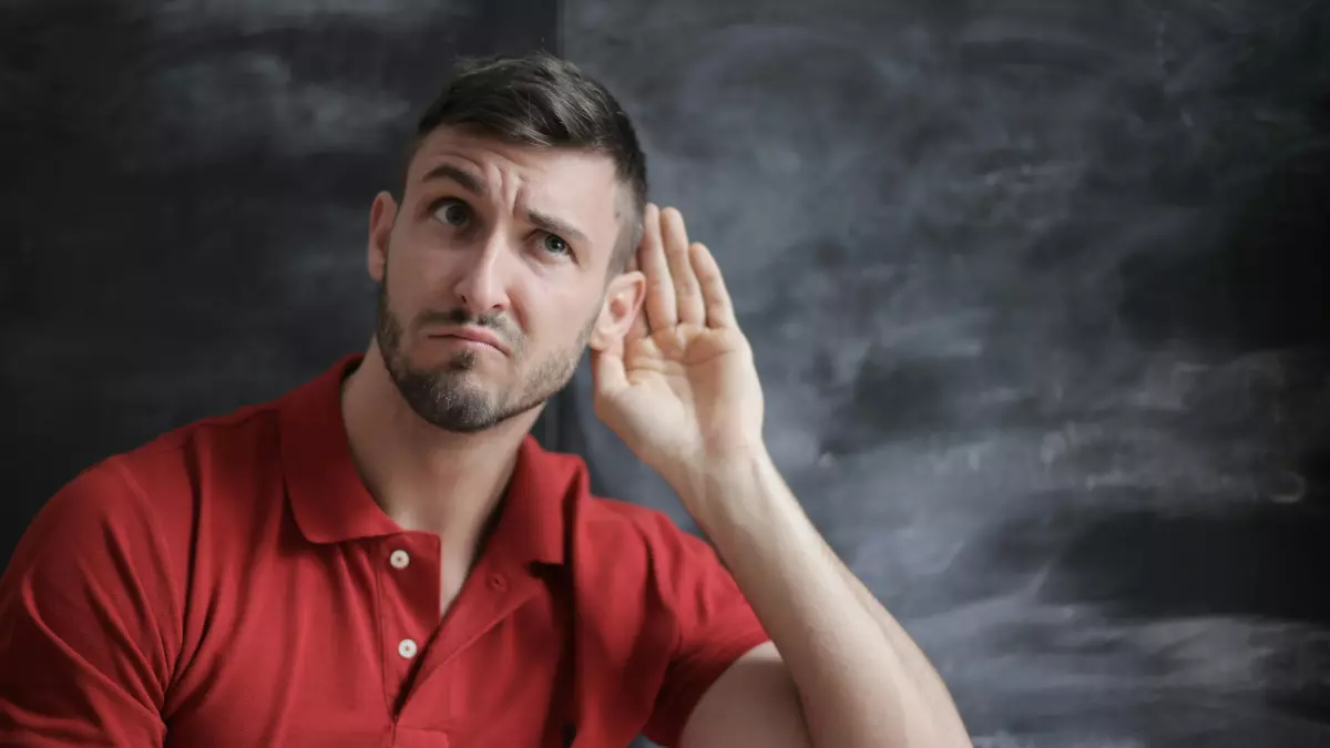 A man with short brown hair is wearing a red polo shirt and is listening intently. He is holding his hand to his ear, seemingly curious to hear what is being said.