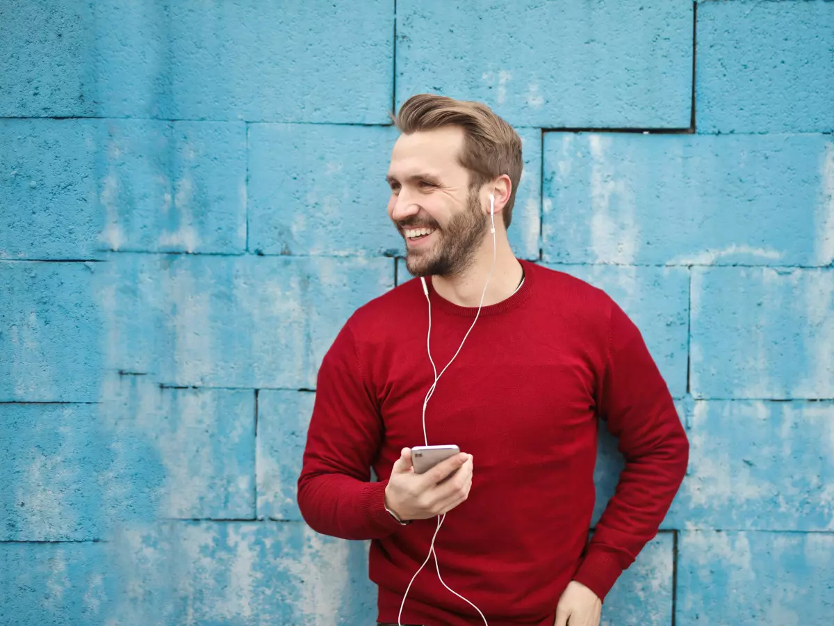 A man with a beard and a red sweater stands in front of a blue wall, smiling as he looks to the right. He is holding a smartphone in his left hand and his right hand is on the phone's cable.