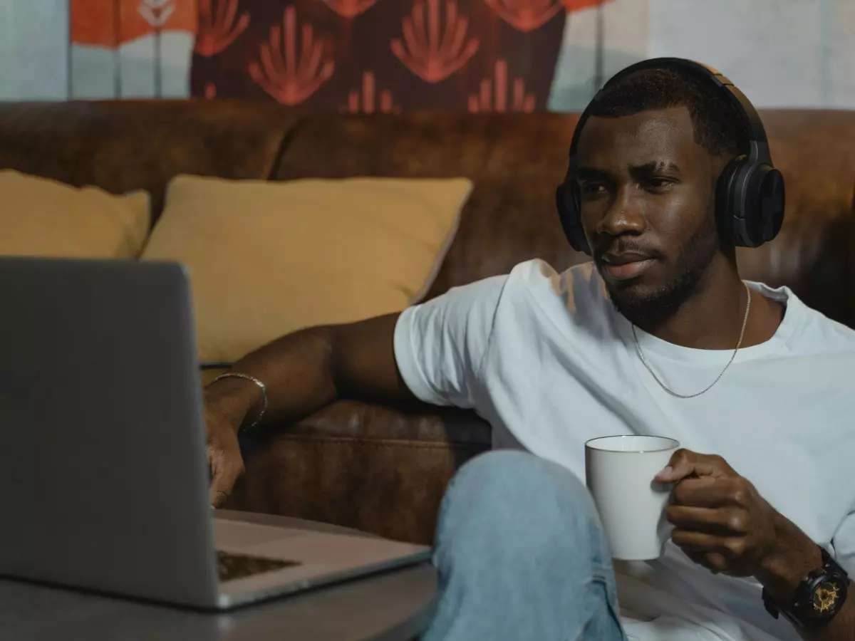 A young man in a white T-shirt and blue jeans is sitting on a brown couch. He is wearing headphones and looking at a laptop in his lap. He is holding a white coffee mug in his right hand.