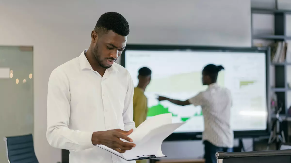 A man in a white shirt is looking down at a document in his hands. He is standing in an office with a large screen in the background, and two other people are working on a project on the screen. 