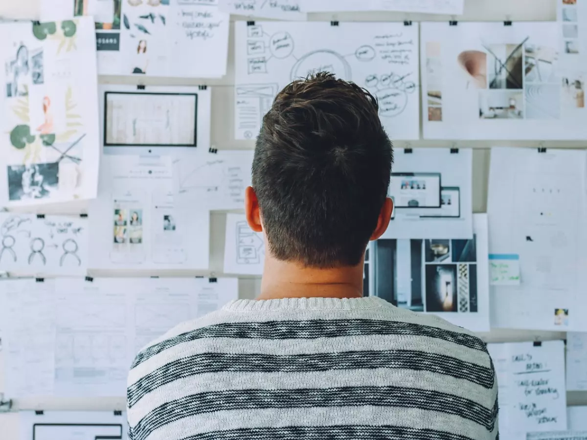 A man standing in front of a wall covered in papers and diagrams, looking at something with a thoughtful expression.