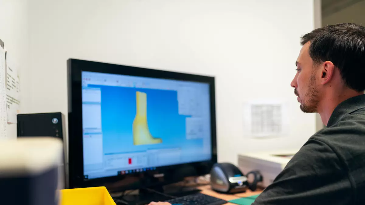 A man is sitting at a desk in front of a computer. He is looking at the screen and appears to be concentrating on his work. The image is shot from a slightly elevated angle, giving the viewer a good view of the man